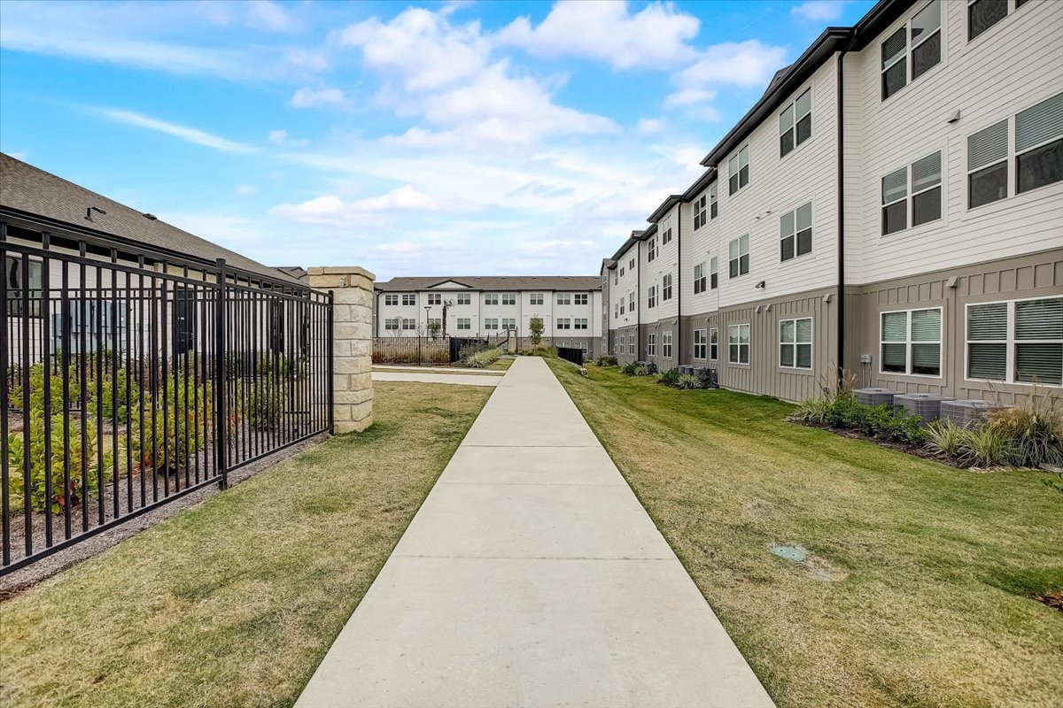 Wide sidewalk between apartment buildings, bordered by lawns, shrubs, and a secure perimeter fence.